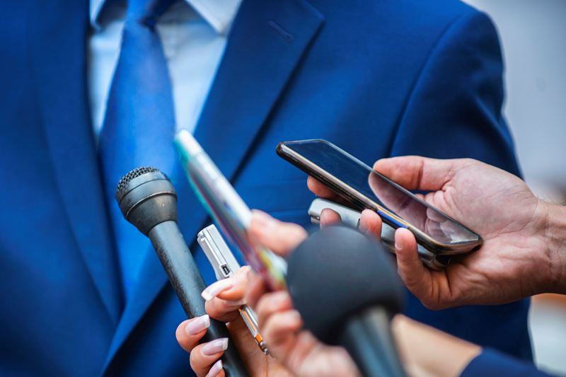 Des journalistes munis de microphones interrogent un homme d’affaires habillé formellement.